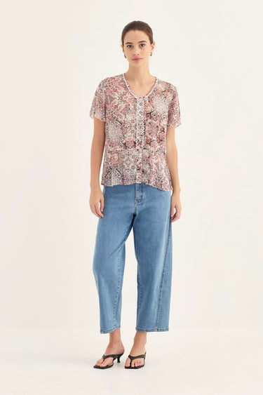 A woman modeling a short-sleeve button-front blouse with an intricate floral and paisley pattern in dusty pink, beige, and brown, paired with light-wash blue cropped jeans by Verge and black sandals, against a plain off-white studio background.