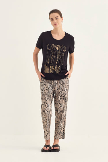 A woman modeling a black short-sleeve t-shirt with a gold and black abstract graphic print on the front, paired with beige and brown snakeskin print pants by Verge, against a plain white studio background.