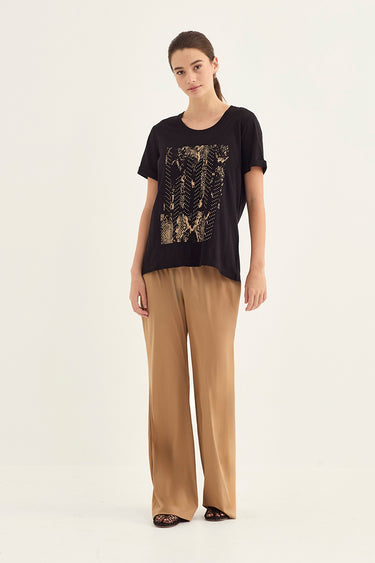 A woman modeling a black short-sleeve t-shirt with a gold-toned graphic print on the front, paired with wide-leg tan pants by Verge, against a plain off-white studio background.