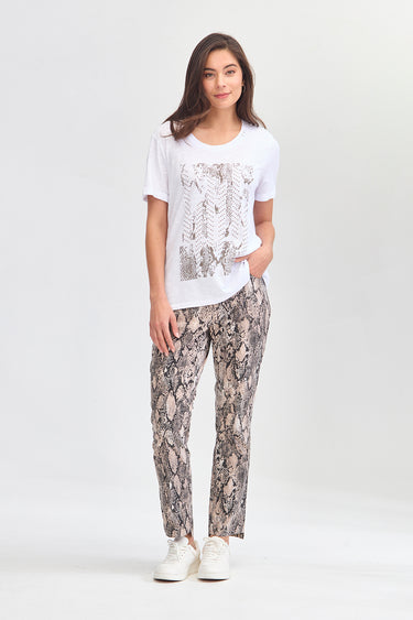 A woman modeling a white short-sleeve t-shirt with a gray leaf pattern and beige snakeskin print pants by Verge, paired with white sneakers, against a light gray studio background.