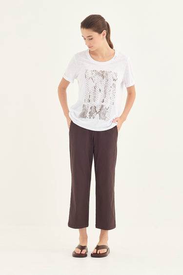 A woman modeling a white short-sleeve t-shirt with a gray abstract graphic print on the front, paired with dark brown cropped pants by Verge and brown slide sandals, against a plain white studio background.