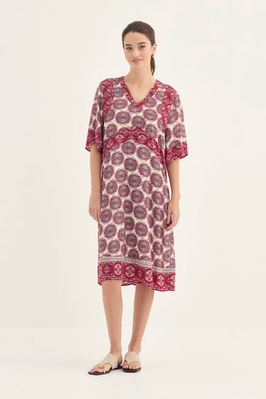 A woman modeling a short-sleeve V-neck kaftan dress by Verge with a red and purple circular pattern on a cream background, set against a plain off-white studio backdrop.