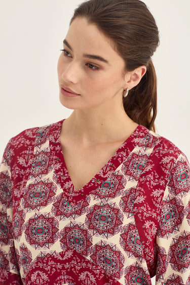 A woman wearing a red and cream patterned V-neck top by Verge, shown from the chest up against a plain light background.