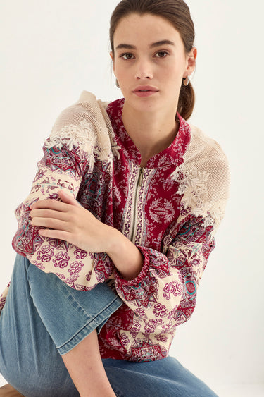 A woman wearing a bohemian-style long-sleeve blouse with a red and cream paisley print, lace trim, and a cream mesh collar, paired with blue jeans by Verge, posed against a light off-white background.