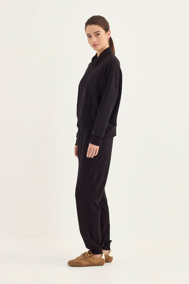 A woman modeling a black long-sleeve top and matching black pants by Verge, paired with brown suede shoes, against a plain off-white studio background.