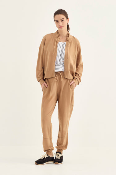 A woman modeling a matching tan two-piece lounge set consisting of a lightweight zip-up jacket and drawstring sweatpants, worn over a white and navy striped shirt by Verge with black sneakers, against a plain off-white studio background.