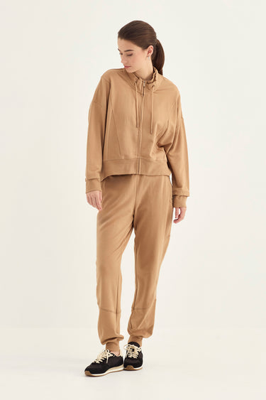 A woman modeling a matching tan two-piece tracksuit consisting of a cropped zip-up hoodie and jogger pants by Verge, paired with black sneakers, against a plain off-white studio background.