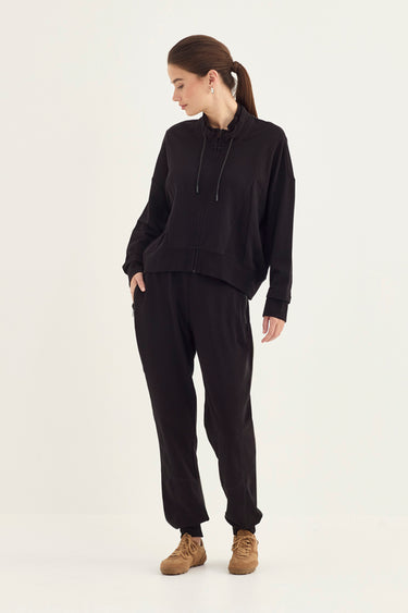 A woman modeling a black two-piece loungewear set consisting of a cropped zip-up hoodie and matching jogger pants by Verge, paired with tan suede boots, against a plain off-white studio background.