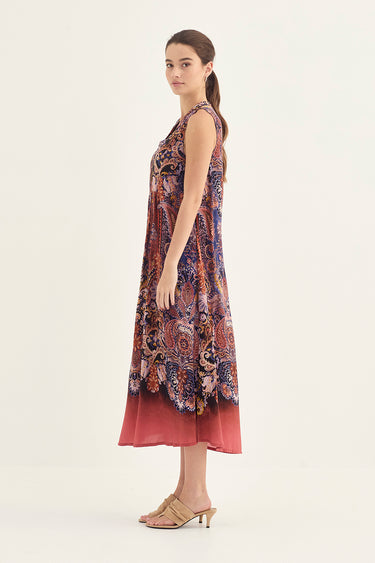 A woman modeling a sleeveless midi dress by Verge with a multicolor paisley pattern in shades of purple, blue, and orange, featuring a solid rust-red sheer overlay at the hem, set against a plain off-white studio background.