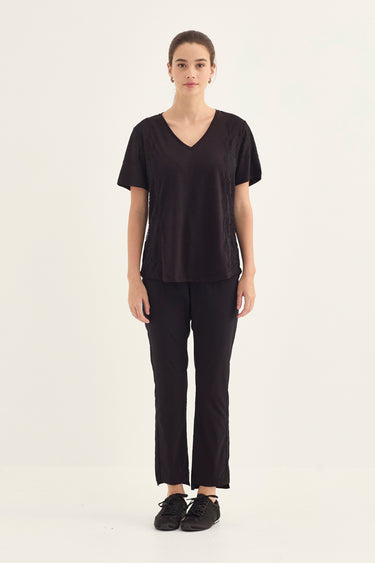 A woman stands against a plain light background wearing a loose black short-sleeve V-neck top, black pants by Verge, and black sneakers.