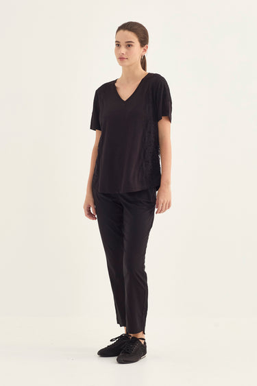 A woman modeling a black short-sleeve V-neck top and matching black pants by Verge with black sneakers, standing against a plain off-white studio background.