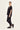 A woman modeling a black short-sleeve V-neck top and matching black pants by Verge with black sneakers, standing in profile against a plain light background.