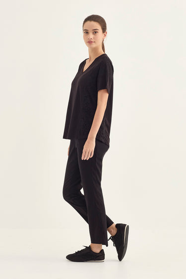 A woman modeling a black short-sleeve V-neck top and matching black pants by Verge with black sneakers, standing in profile against a plain light background.