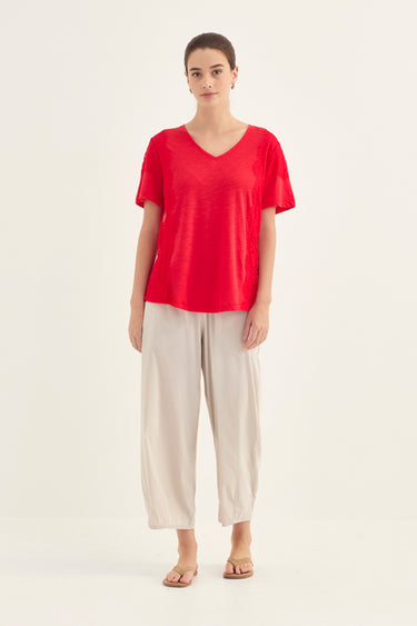A woman modeling a bright red short-sleeve V-neck top paired with light beige wide-leg cropped pants by Verge and flat sandals, standing against a plain off-white studio background.