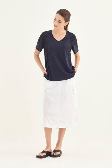 A woman modeling a short-sleeve navy blue V-neck top and a white midi skirt by Verge, paired with black slide sandals, against a plain off-white studio background.