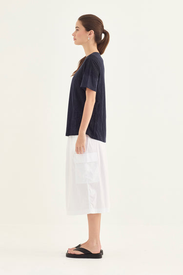 A woman shown in side profile wearing a dark navy short-sleeve top, a white midi skirt with large front patch pockets by Verge, and black slide sandals against a plain light background.