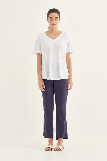 A woman modeling a white short-sleeve V-neck top and navy blue cropped pants by Verge against a plain off-white studio background.