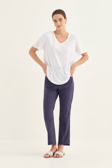 A woman modeling a white short-sleeve V-neck top and navy blue cropped pants by Verge against a plain off-white studio background.