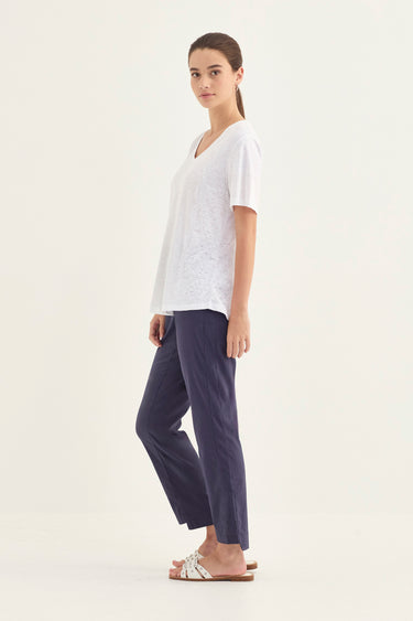 A woman modeling a white short-sleeve V-neck top and navy blue cropped pants by Verge, standing in a side profile against a plain off-white studio background.