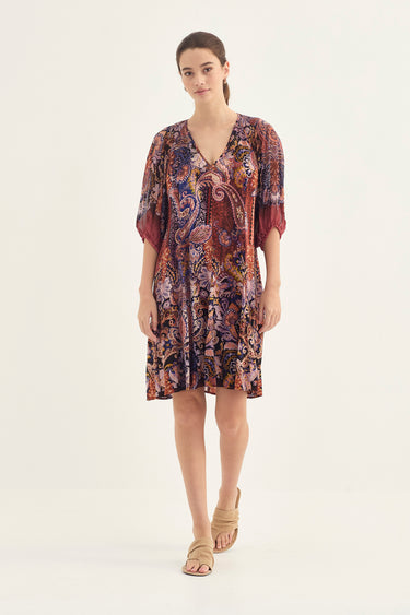 A woman modeling a short-sleeve V-neck dress by Verge with a dense paisley and floral print in burgundy, purple, and brown tones, paired with beige flat sandals, against a plain off-white studio background.