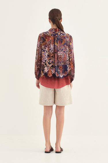 A woman viewed from the back wearing a long-sleeve button-up shirt by Verge with a dense paisley and floral pattern in purple, orange, brown, and blue tones, paired with light beige shorts and black sandals, against a plain off-white studio background.