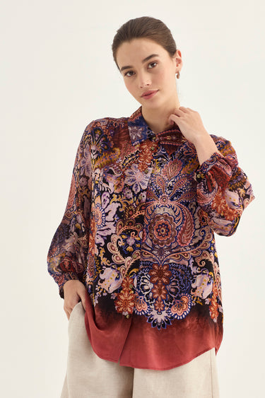 A woman modeling a long-sleeve button-up blouse with an intricate multicolor paisley and floral print in red, blue, purple, and gold tones, paired with light beige trousers by Verge, against a plain off-white studio background.