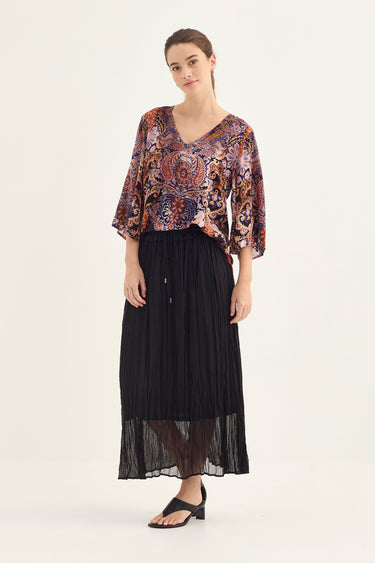 A woman modeling a bohemian-style patterned blouse with three-quarter sleeves and a long black pleated skirt by Verge, standing against a plain off-white studio background.