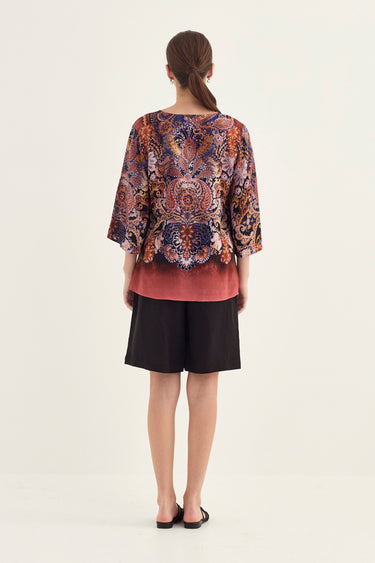 A woman viewed from the back wearing a loose short-sleeve top with a multicolor paisley pattern and a dusty rose gradient hem, paired with black shorts by Verge, against a plain light background.