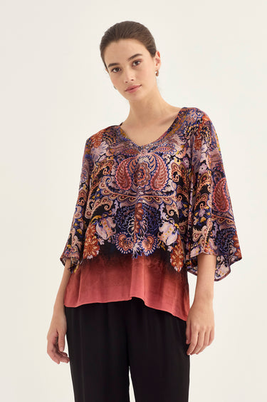 A woman modeling a loose, short-sleeved V-neck top by Verge with a multicolor paisley and floral print on the upper portion and a solid dusty rose gradient on the lower portion, paired with black pants against a plain light background.