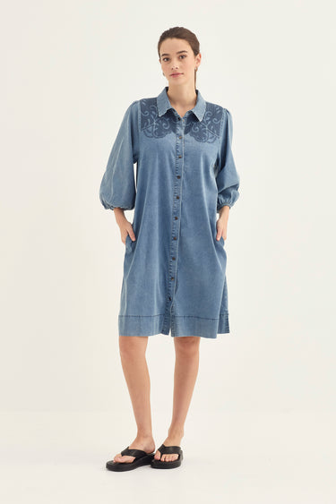 A woman modeling a light blue denim shirt dress by Verge with dark blue floral embroidery across the chest, paired with black slide sandals, against a plain off-white studio background.