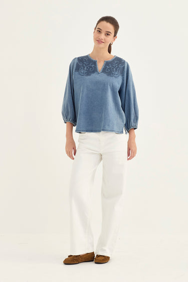 A woman modeling a light blue denim long-sleeve blouse with sleeves and white wide-leg jeans by Verge, standing against a plain white studio background.