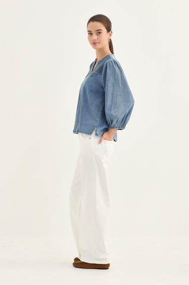 A woman modeling a light blue denim long-sleeve blouse with voluminous puff sleeves and white wide-leg jeans by Verge, standing against a plain white studio background.