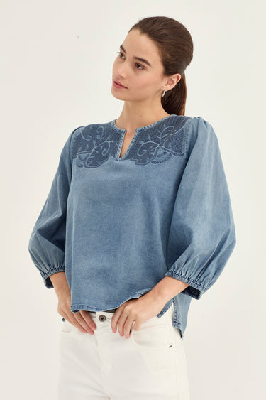 A woman wearing a light blue denim long-sleeve blouse with a dark blue embroidered yoke and white pants, photographed against a plain off-white studio background.