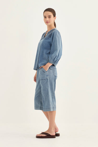 A woman modeling a light blue denim two-piece set consisting of a long-sleeve button-up shirt and matching cropped shorts by Verge, paired with dark brown flip-flop sandals, against a plain off-white studio background.