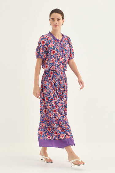 A woman modeling a short-sleeved blouse and a skirt by Verge with a purple, red, and blue floral pattern set against a plain white studio background.