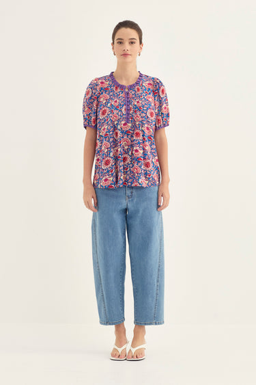 A woman stands against a plain light background wearing a short-sleeve floral blouse with blue wide-leg cropped jeans by Verge and white sandals.