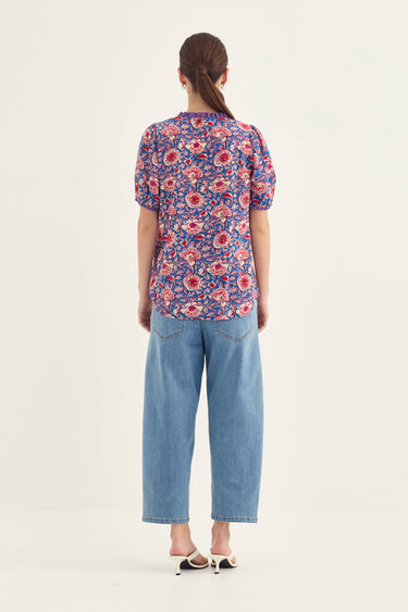 A woman viewed from the back wearing a short-sleeve floral blouse with a purple, red, and blue pattern, paired with light blue cropped jeans by Verge and white pointed-toe flats, against a plain off-white studio background.