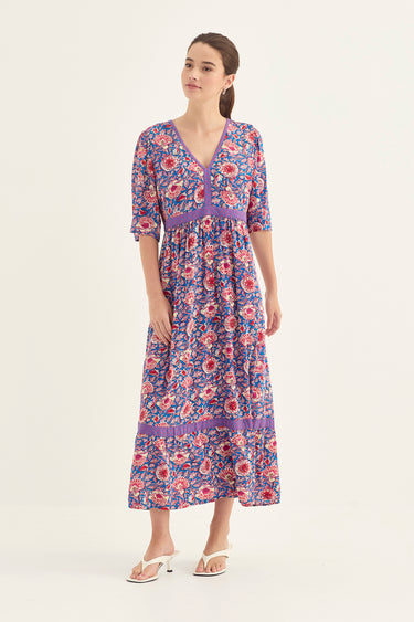 A woman modeling a purple floral dress by Verge with three-quarter sleeves and a V-neckline, paired with white heeled sandals, against a plain off-white studio background.