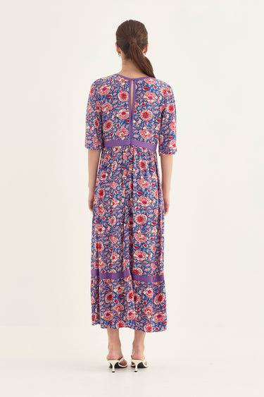 A woman viewed from the back wearing a knee-length floral dress by Verge with three-quarter sleeves and a purple waistband, paired with white heeled sandals, against a plain light background.