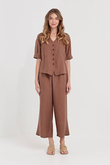 A woman modeling a matching bronze two-piece set consisting of a short-sleeve button-front top and wide-leg cropped pants by Verge, paired with beige flat sandals, against a plain light gray studio background.