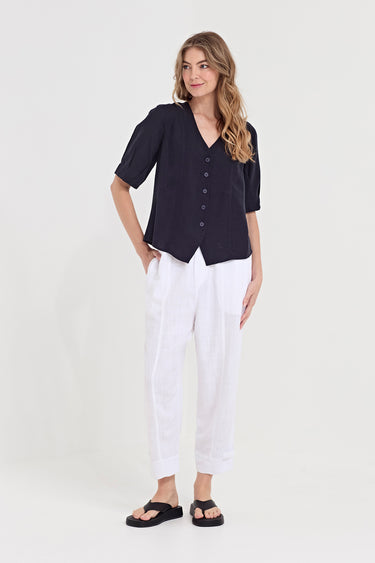 A woman modeling a short-sleeve dark navy button-front blouse with a V-neck and pleated details, paired with white trousers by Verge and black sandals, against a plain light gray studio background.