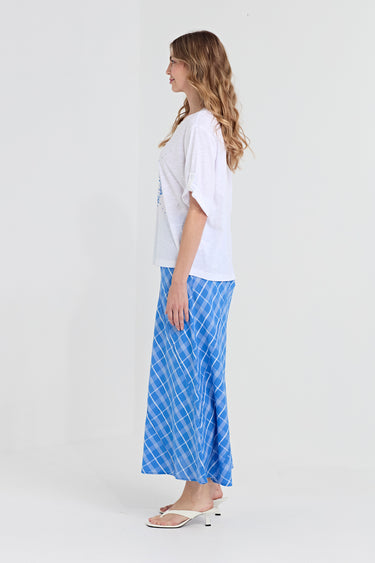 A woman stands in profile wearing a loose white short-sleeve top and a long blue skirt with a white diagonal grid pattern by Verge, paired with white heeled sandals, against a plain light gray studio background.