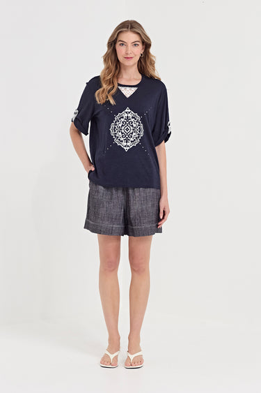 A woman modeling a navy blue short-sleeve t-shirt with a white mandala print on the front, paired with gray patterned shorts by Verge and white flip-flop sandals, against a plain light gray background.