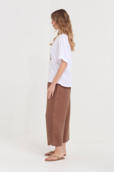 A woman stands in profile wearing a loose white short-sleeve T-shirt and brown wide-leg cropped pants by Verge, paired with tan sandals, against a plain light gray studio background.