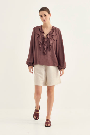 A woman modeling a brown ruffled long-sleeve blouse with cream shorts and brown sandals by Verge, standing against a plain off-white studio background.