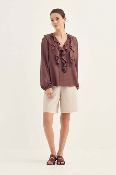 A woman modeling a long-sleeve ruffled blouse in a muted mauve-brown color, paired with light beige shorts and dark brown slide sandals by Verge, against a plain off-white studio background.