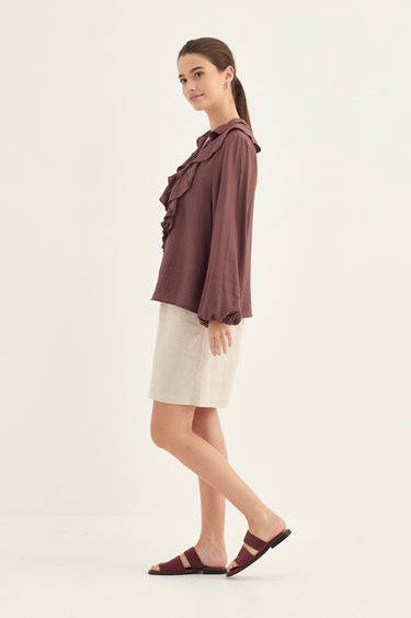 A woman modeling a dark brown ruffled long-sleeve blouse with a high collar, paired with a light beige skirt by Verge and dark brown slide sandals, set against a plain off-white studio background.