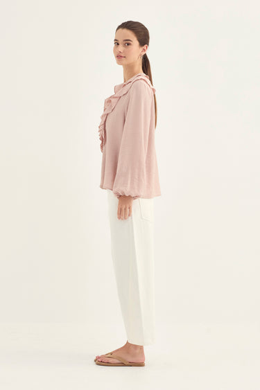 A woman stands in profile wearing a light pink long-sleeve blouse with ruffled detailing and white wide-leg pants by Verge, against a plain off-white studio background.