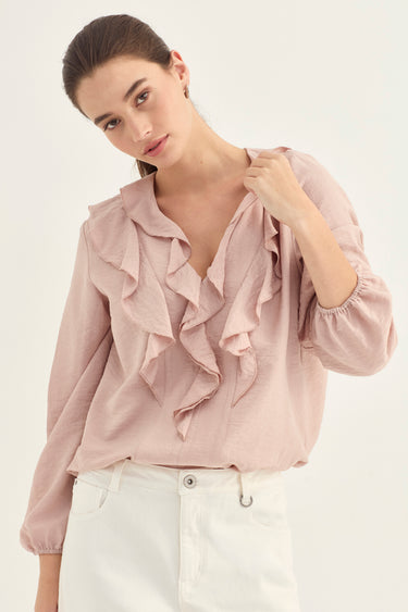 A woman wearing a dusty pink ruffled blouse with long sleeves and white pants by Verge, photographed against a plain light background.