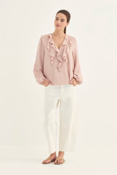 A woman modeling a light pink long-sleeve blouse with layered ruffles along the V-neckline, paired with white wide-leg cropped pants and flat sandals by Verge, against a plain white studio background.